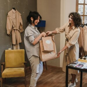 Two women exchanging shopping bags in a stylish boutique, showcasing fashion and smiles.